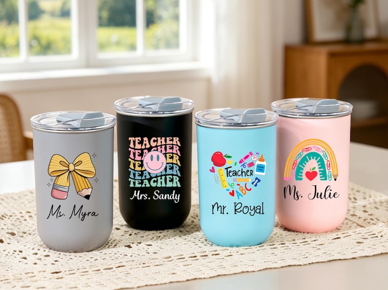 May include: Four cylindrical tumblers with clear lids, each featuring unique teacher-themed graphics and names. The gray tumbler shows a pencil bow, the black one has a smiley face, the light blue one displays school supplies, and the pink one a rainbow.