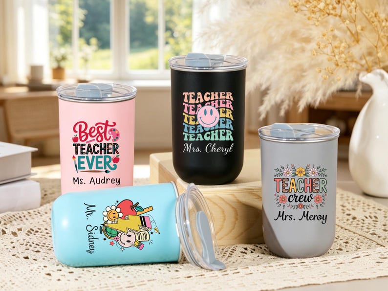 May include: Four stainless steel tumblers with clear lids. One pink tumbler reads Best Teacher Ever Ms. Audrey. One black tumbler features a smiley face and the text Teacher Mrs. Cheryl. One gray tumbler says Teacher crew Mrs. Meroy. One light blue tumbler says Mr. Sidney.