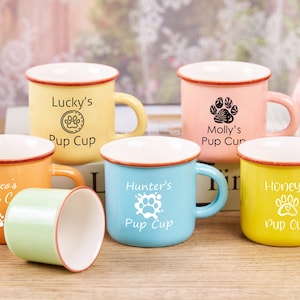 May include: Five colorful ceramic mugs with paw print designs and personalized names. The mugs are yellow, orange, light blue, pink, and green. Each mug has the text "Pup Cup" and a name, including "Coco's", "Lucky's", "Molly's", "Hunter's", and "Honey's".