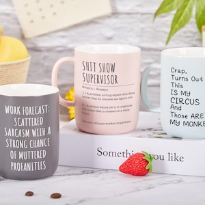 May include: Three ceramic mugs with humorous text. One is gray with "Work Forecast: Scattered Sarcasm...", another is pink with "Shit Show Supervisor", and the third is light blue with "Crap. Turns Out This Is My Circus...". A strawberry and fruit are in the background.