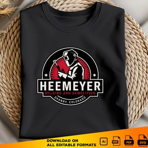 May include: Black t-shirt with a red, white, and black logo featuring a welder with a welding torch. The logo says "Heemeyer Welding and Demolition Granby, Colorado".