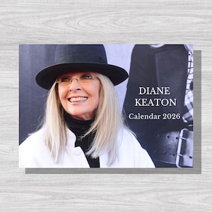 Puede incluir: Calendario con una mujer sonriente con sombrero negro y gafas. El texto "DIANE KEATON Calendar 2026" se muestra en el lado derecho de la imagen. Fondo de superficie de madera gris claro.