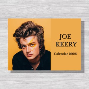 以下が含まれることがあります： ジョー・キーリーの肖像画が描かれたカレンダー。顔には黄色の稲妻のデザインが施されています。カレンダーはマスタードイエローの背景で、「JOE KEERY Calendar 2026」の文字が書かれています。