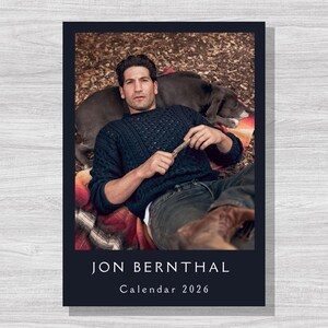 Può includere: Calendario con bordo blu scuro, raffigurante un uomo con maglione blu navy e jeans, sdraiato su una coperta con un cane. Il testo "JON BERNTHAL Calendar 2026" è bianco.