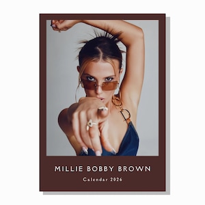 Puede incluir: Calendario con borde marrón que presenta un retrato de Millie Bobby Brown. Lleva gafas de sol y señala al espectador. El texto "MILLIE BOBBY BROWN Calendar 2026" está impreso en la parte inferior.