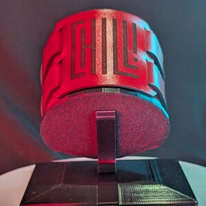 Puede incluir: Un objeto rojo y negro impreso en 3D con un diseño geométrico, montado sobre una base blanca y negra. La base se encuentra sobre una plataforma giratoria blanca, iluminada por luces rojas y azules.