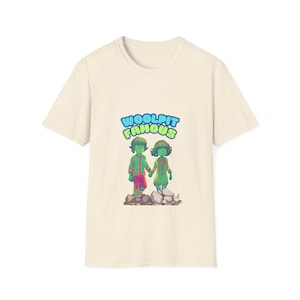 Puede incluir: Una camiseta de color beige claro con un estampado gráfico de dos niños de dibujos animados verdes que se toman de la mano. El texto "Woolpit Famous" está impreso en azul y verde sobre los niños.