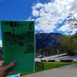 Può includere: Un libretto verde con il testo "The Happy 100's" e un'illustrazione in bianco e nero di un albero. Il libretto è tenuto davanti a un cielo blu con nuvole bianche e una catena montuosa sullo sfondo.