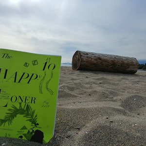 Puede incluir: Un libro verde con el título "The Happy Loner" y el número "10" en la portada. El libro está tirado en una playa de arena con un tronco grande en el fondo.
