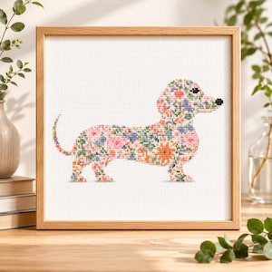 Peut inclure: Une broderie au point de croix encadrée représentant un chien teckel avec un motif floral rose, bleu, orange et vert. Le chien est sur fond blanc, encadré dans un cadre en bois brun clair. L'œuvre est sur une surface en bois.