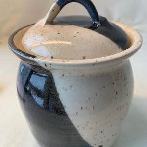 Puede incluir: Un tarro de cerámica para ajo con tapa y asa. El tarro es una mezcla de blanco y azul oscuro, con un patrón moteado. La tapa tiene un asa azul oscuro. El tarro tiene varios agujeros cerca de la parte inferior.