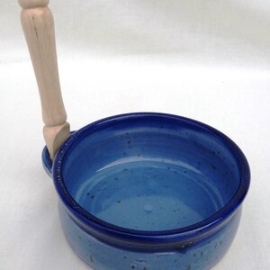 May include: A blue ceramic bowl with a wooden handle. The bowl has a speckled glaze and a dark blue rim.