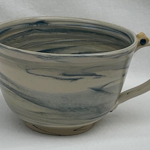 Peut inclure: Une tasse en céramique avec un large bol et une anse. La tasse présente un motif marbré dans les tons de blanc, de gris et de bleu. La poignée a un petit accent en bois avec un point bleu. La tasse repose sur une petite base ronde.