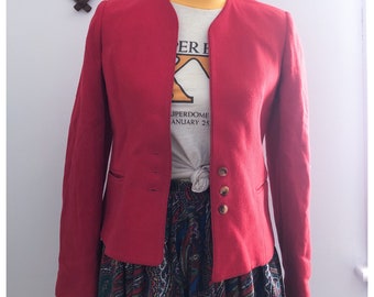 May include: A red blazer with a mandarin collar and two gold buttons. The blazer is worn over a white t-shirt with the text "Superdome January 25" printed on it.