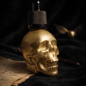 May include: A gold-colored skull-shaped bottle with a black dropper top. The bottle is set against a dark background, with a wand and a golden winged object in the background. The skull has detailed features.