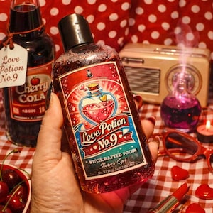 May include: A bottle of "Love Potion No. 9" shower gel with a heart design is held. A bottle of Cherry Cola, cherries, lipstick, and heart decorations are also visible. The background features a red and white polka dot pattern.