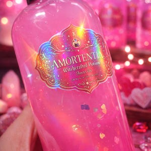 Puede incluir: Una botella rosa de gel de ducha "Amortentia Witchcrafted Potion". La etiqueta tiene un diseño dorado e iridiscente. El líquido del interior es rosa y contiene purpurina. La botella tiene una tapa negra.