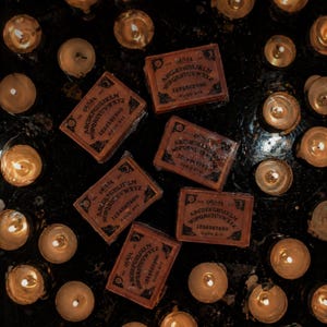 Puede incluir: Vista cenital de varias velas encendidas que rodean pequeñas tablas Ouija rectangulares. Las tablas son marrones con letras negras y presentan el alfabeto, números y las palabras "Ouija" y "Good Bye". Las velas son el foco, sobre un fondo oscuro.