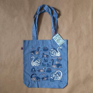 Puede incluir: Una bolsa de tela azul claro con un estampado blanco de cisnes, gansos y patos sobre un fondo azul. La bolsa tiene una etiqueta blanca que dice "100% algodón tote".
