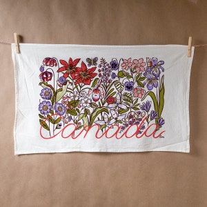 Puede incluir: Toalla de cocina blanca con un diseño floral colorido que presenta lirios rojos, flores moradas y blancas y hojas verdes. La palabra "Canadá" está escrita en cursiva roja en la parte inferior de la toalla.