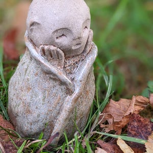 May include: A small, stone sculpture of a serene figure, possibly a monk. The figure is seated, with hands resting on the face. The sculpture has a textured, weathered appearance, and is set amidst green grass and fallen leaves. The sculpture is likely made of stone.