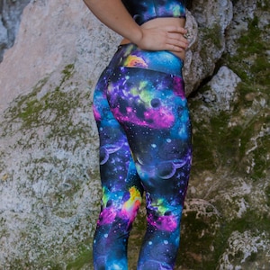 Puede incluir: Un par de leggings negras con un estampado de galaxia colorido. Las leggings presentan una variedad de colores, incluyendo rosa, azul, verde y amarillo. El estampado recuerda a un cielo estrellado.