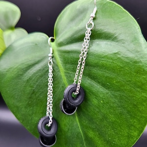 May include: A pair of dangle earrings with silver chains and black rubber tire-shaped charms. The earrings are hanging from a green leaf.