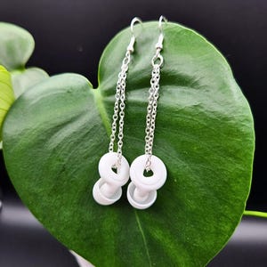 May include: White ceramic circle earrings with silver chains hanging from the bottom. The earrings are displayed on a green leaf.