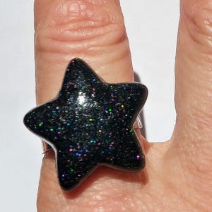 Puede incluir: Un anillo negro en forma de estrella con purpurina iridiscente incrustada en la resina. El anillo se lleva en un dedo.