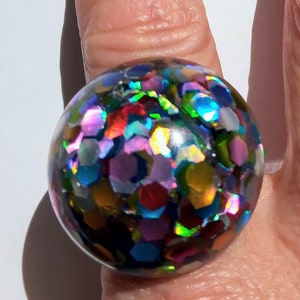 May include: A large, round ring with a clear resin dome filled with iridescent, multi-colored glitter. The glitter reflects hues of blue, green, purple, and gold. The ring is set on a silver band.