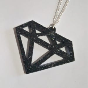 Può includere: Collana con pendente a forma di diamante nero glitterato. Il pendente ha un design geometrico ed è sospeso a una catena d'argento. La forma del diamante è aperta, con sfaccettature visibili. I glitter aggiungono brillantezza.