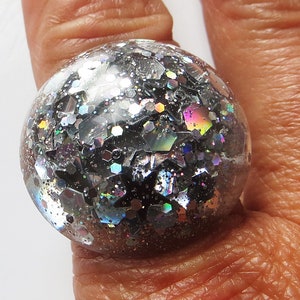 May include: A large, round ring featuring a clear orb filled with silver and black glitter, with iridescent flashes. The ring is worn on a finger, highlighting the sparkly design. The orb shape of the ring is the main feature.