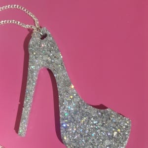 May include: A silver glitter high-heeled shoe pendant on a silver chain. The pendant is shaped like a stiletto heel and is covered in sparkling glitter. The background is a solid pink color.
