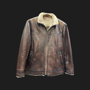 May include: A brown leather jacket with a beige shearling collar and lining. The jacket features a full zipper closure, two front pockets, and decorative stitching along the seams. The jacket is displayed against a black background.