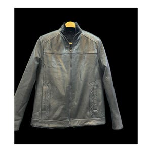 Puede incluir: Chaqueta de cuero negra con cremallera completa, cuello alzado y dos bolsillos delanteros. La chaqueta está colgada de una percha de madera sobre un fondo negro. La chaqueta parece ser de talla mediana.