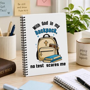 Puede incluir: Un cuaderno de espiral blanco con el texto "With God in my backpack, no test scares me" y una ilustración de una mochila, libros y una cruz. Encuadernación en espiral negra.