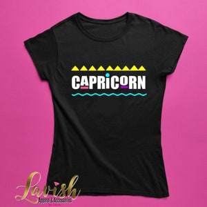 May include: Black t-shirt with a graphic design featuring the word "CAPRICORN" in colorful letters with a blue wavy line and yellow triangles above the word.