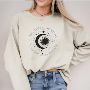 Könnte beinhalten: Ein hellbeiger Pullover mit einem schwarzen Grafikdesign einer Sonne und eines Mondes mit dem Text "Glaube an die Sterne, umarme deine Zukunft".