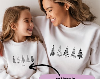May include: White Christmas sweaters with a black design of stylized Christmas trees. The smaller sweater has the word "MINI" printed below the trees. The larger sweater has the text "optionale Personalisierung" printed below the trees.