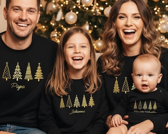 May include: A family wearing matching black Christmas sweaters with gold Christmas tree designs. The sweaters have the names "Papa" and "Johanna" printed on them. The baby is wearing a black onesie with the text "Mein erster Weihnachten".
