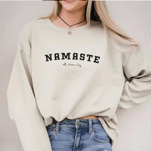 Könnte beinhalten: Ein hellbeiger Pullover mit dem Aufdruck "NAMASTE all damn day" in Schwarz.