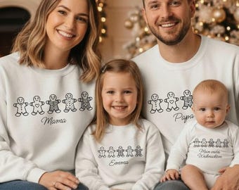 May include: A family wearing matching white sweatshirts and a onesie. Each top features a line of gingerbread people and a name: Mama, Papa, Emma, and the baby's onesie says "Meine erstes Weihnachten". The family is smiling.
