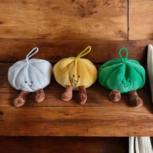 May include: Three plush toys shaped like rounded, pleated dumplings in white, yellow, and green. Each toy features a small loop at the top, two brown dangling legs, and a simple embroidered face with two eyes and a curved mouth line.