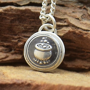 May include: A silver-toned pendant necklace featuring a round charm with a cauldron design. The cauldron is depicted with bubbles rising from it. The necklace chain is also silver and hangs against a textured, brown background.
