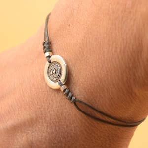 Può includere: Un braccialetto in corda grigia con un ciondolo circolare argentato con un design dettagliato. Il ciondolo è centrato sul braccialetto.