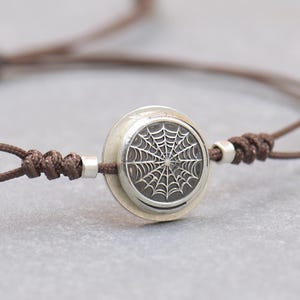 Op de afbeelding: Bruin koordarmband met een zilverkleurige bedel met een gedetailleerd spinnenwebontwerp. De verstelbare armband heeft kleine zilveren kralen aan de uiteinden en een schuifknoopsluiting. De bedel is cirkelvormig.
