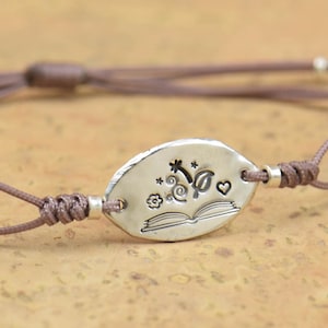 Puede incluir: Un brazalete de cordón marrón con un dije de plata que representa un libro abierto con un corazón, una estrella y otros elementos decorativos.
