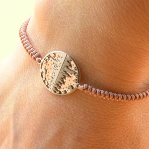 May include: A brown cord bracelet with a round metal charm featuring a tree design.