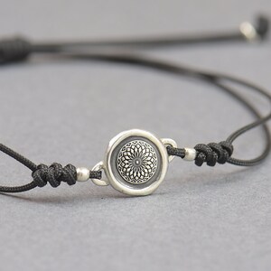 Può includere: Braccialetto con cordino nero e elemento centrale circolare argentato. L'elemento centrale presenta un design simmetrico dettagliato. Il braccialetto è regolabile con nodi scorrevoli e piccole perline argentate alle estremità.