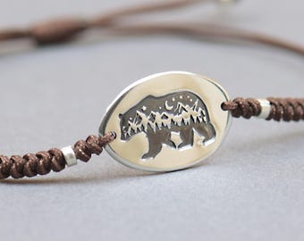 May include: A brown cord bracelet with a silver oval pendant. The pendant features a bear silhouette with a mountain and moon design. The bracelet has adjustable sliding knots and small silver beads.